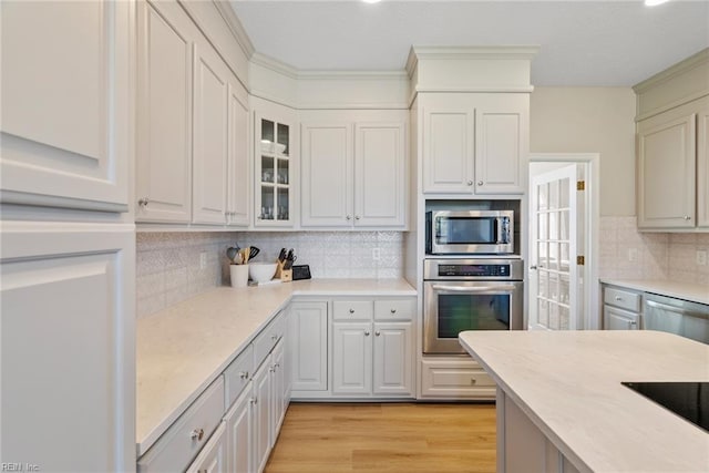 kitchen featuring light countertops, light wood finished floors, stainless steel appliances, tasteful backsplash, and glass insert cabinets