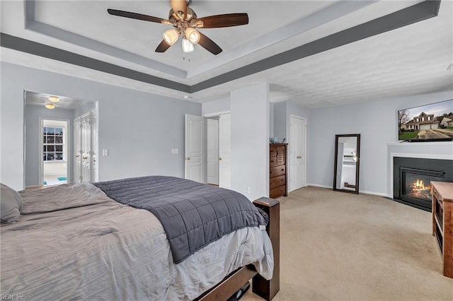 bedroom featuring ceiling fan, baseboards, a tray ceiling, light carpet, and a glass covered fireplace