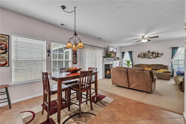 dining room featuring light tile patterned floors, visible vents, ceiling fan with notable chandelier, a warm lit fireplace, and light colored carpet