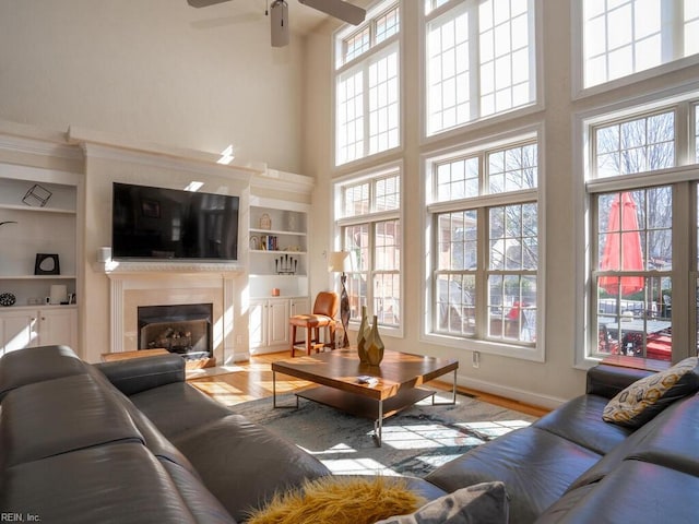 living room with a wealth of natural light, built in shelves, a fireplace with flush hearth, wood finished floors, and ceiling fan