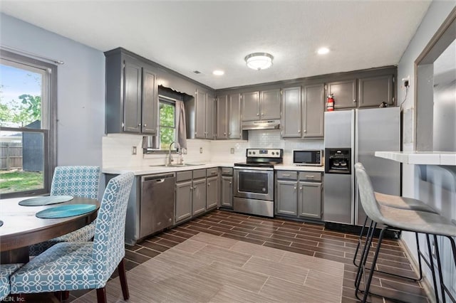 kitchen featuring decorative backsplash, gray cabinets, a sink, under cabinet range hood, and stainless steel appliances