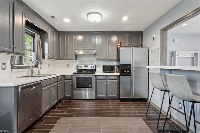 kitchen with under cabinet range hood, tasteful backsplash, a sink, gray cabinetry, and stainless steel appliances