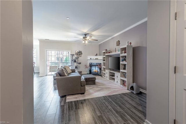 living area with a glass covered fireplace, crown molding, baseboards, ceiling fan, and dark wood-style flooring