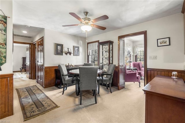 dining space featuring light carpet, wooden walls, a ceiling fan, and a wainscoted wall