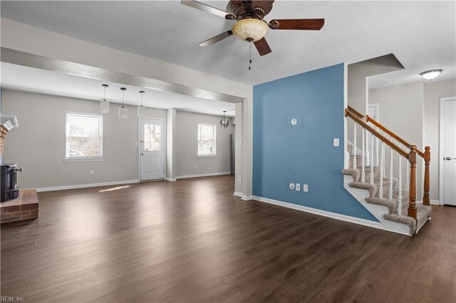 unfurnished living room featuring dark wood-style flooring, baseboards, stairway, a ceiling fan, and a wood stove