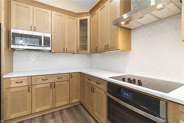 kitchen with glass insert cabinets, wall chimney range hood, light countertops, dark wood finished floors, and appliances with stainless steel finishes