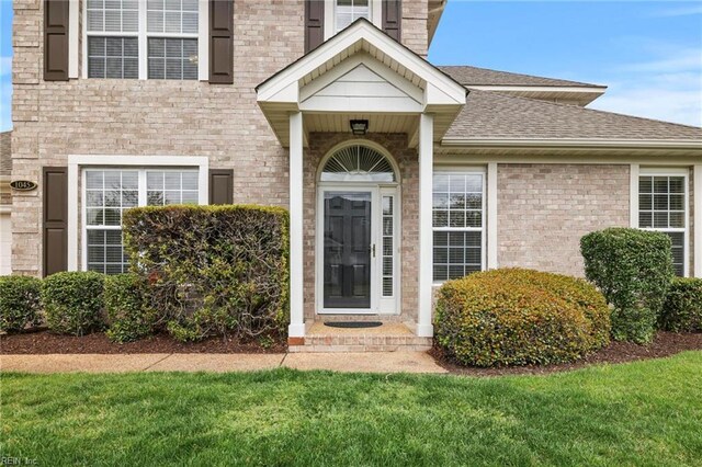 doorway to property featuring a yard, brick siding, and roof with shingles