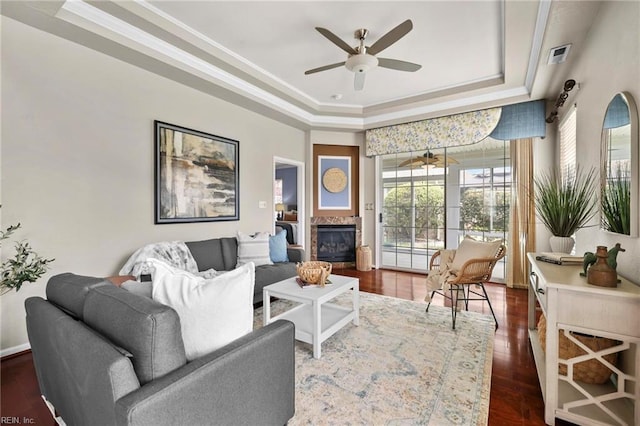 living room with ceiling fan, ornamental molding, dark wood finished floors, a raised ceiling, and a glass covered fireplace