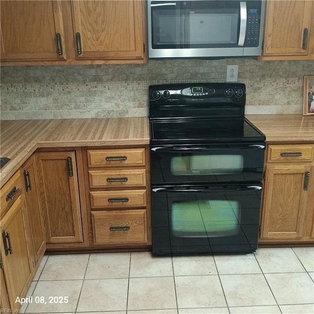 kitchen featuring light tile patterned flooring, light countertops, stainless steel microwave, black electric range, and backsplash