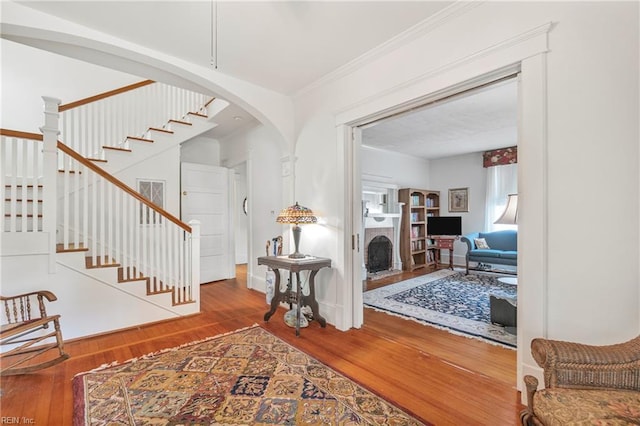 foyer entrance with crown molding, arched walkways, a fireplace with raised hearth, stairway, and wood finished floors