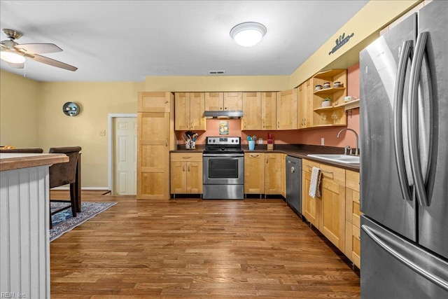 kitchen featuring light brown cabinetry, appliances with stainless steel finishes, under cabinet range hood, a sink, and dark countertops