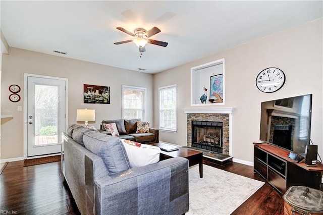 living room with ceiling fan, visible vents, a stone fireplace, baseboards, and dark wood-style floors
