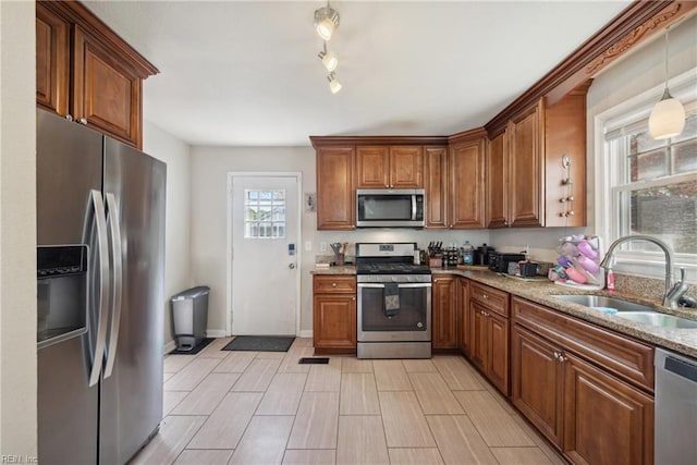 kitchen featuring brown cabinetry, appliances with stainless steel finishes, light stone counters, a sink, and decorative light fixtures