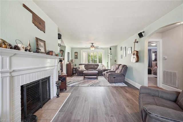 living room with light wood-style floors, arched walkways, a ceiling fan, visible vents, and a brick fireplace