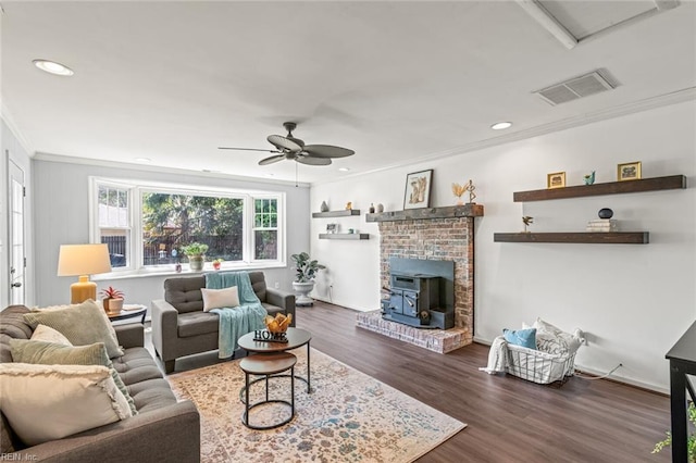 living room with a wood stove, a ceiling fan, ornamental molding, visible vents, and wood finished floors