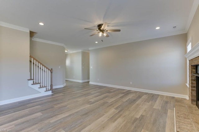 unfurnished living room with a brick fireplace, ornamental molding, ceiling fan, light wood-style floors, and stairs