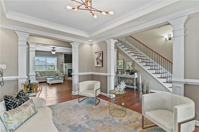 living room featuring a tray ceiling, a glass covered fireplace, ceiling fan with notable chandelier, wood finished floors, and ornamental molding