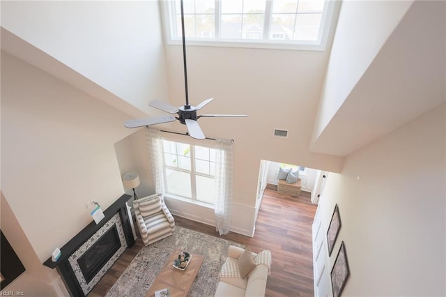 living room with wood finished floors, a glass covered fireplace, ceiling fan, a high ceiling, and visible vents