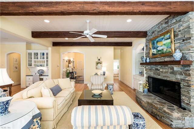 living area with a ceiling fan, light wood-type flooring, arched walkways, a fireplace, and beam ceiling