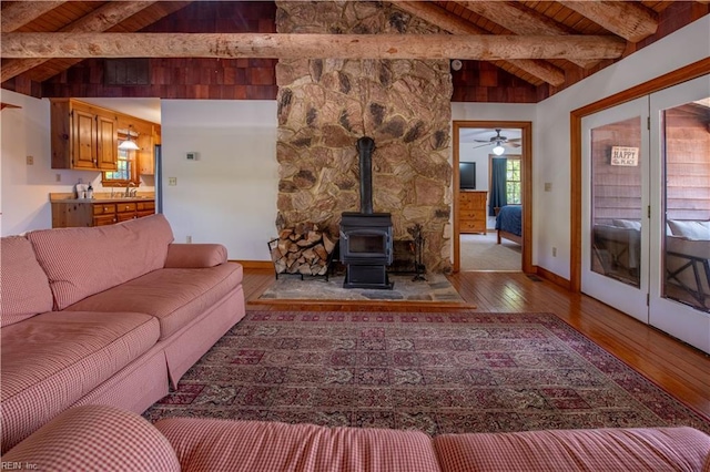 living area with hardwood / wood-style floors, a wood stove, vaulted ceiling with beams, baseboards, and ceiling fan