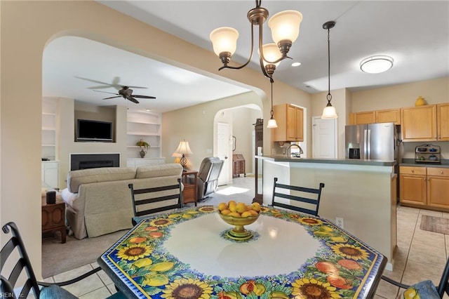 dining space with arched walkways, built in shelves, ceiling fan with notable chandelier, a fireplace, and light tile patterned floors
