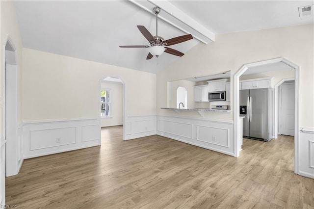 unfurnished living room with a wainscoted wall, arched walkways, lofted ceiling with beams, light wood-style flooring, and ceiling fan