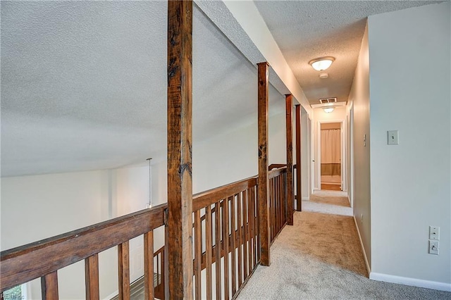 hallway with an upstairs landing, a textured ceiling, light carpet, baseboards, and visible vents