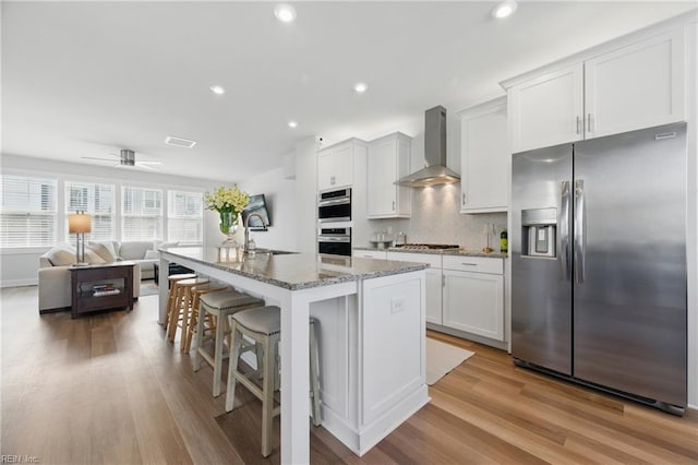 kitchen with decorative backsplash, stainless steel appliances, light wood-style floors, a kitchen breakfast bar, and wall chimney exhaust hood