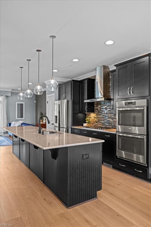 kitchen featuring a sink, light wood-style floors, wall chimney exhaust hood, appliances with stainless steel finishes, and dark cabinets