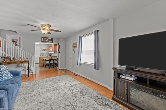 living area featuring stairway, light wood-type flooring, a ceiling fan, and baseboards