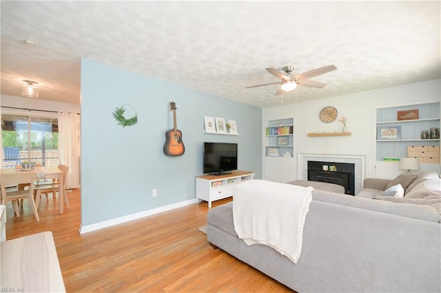 living area featuring light wood-style flooring, built in shelves, ceiling fan, a fireplace, and baseboards