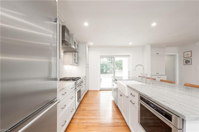 kitchen with a sink, wall chimney exhaust hood, high quality appliances, white cabinets, and an island with sink