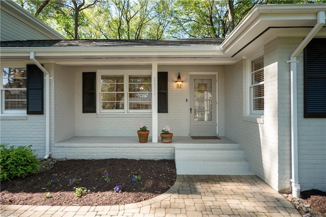 view of exterior entry featuring a shingled roof, covered porch, and brick siding