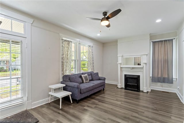 living area featuring a fireplace with flush hearth, ceiling fan, visible vents, baseboards, and dark wood-type flooring