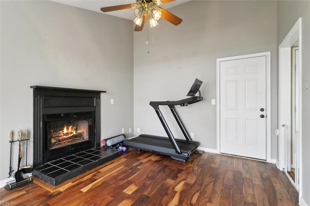 exercise room with a high ceiling, a fireplace, ceiling fan, baseboards, and wood finished floors