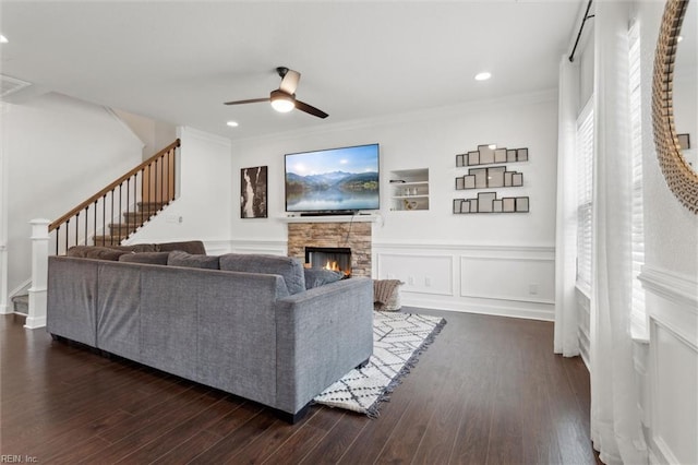 living room with stairs, ornamental molding, a stone fireplace, dark wood-style flooring, and a ceiling fan