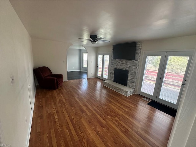 unfurnished living room with ceiling fan, visible vents, arched walkways, wood finished floors, and a stone fireplace