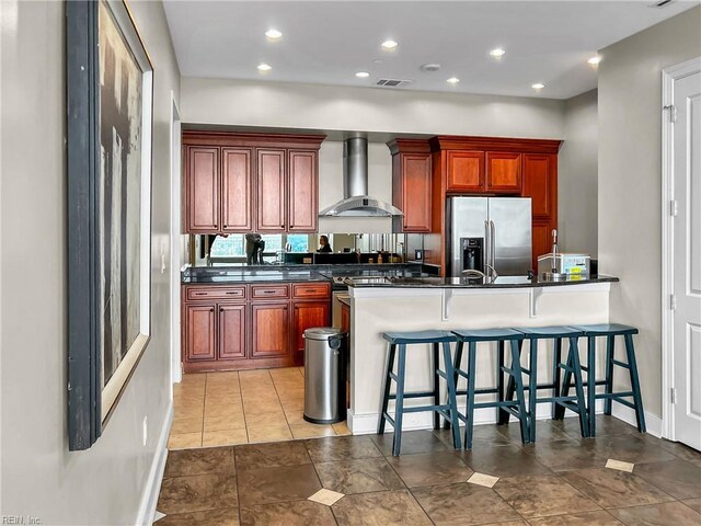 kitchen with a kitchen breakfast bar, stainless steel fridge with ice dispenser, dark countertops, and wall chimney exhaust hood