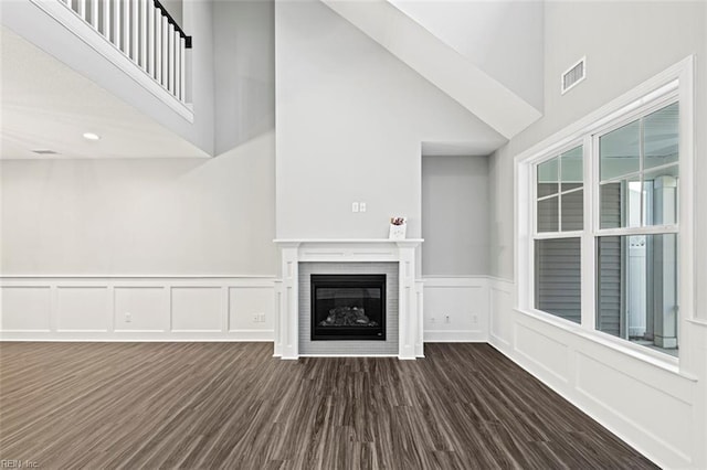 unfurnished living room featuring a wainscoted wall, a glass covered fireplace, dark wood-type flooring, a decorative wall, and a high ceiling