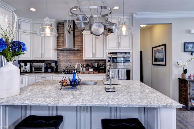 kitchen featuring white cabinets, a kitchen bar, stainless steel double oven, and wall chimney exhaust hood