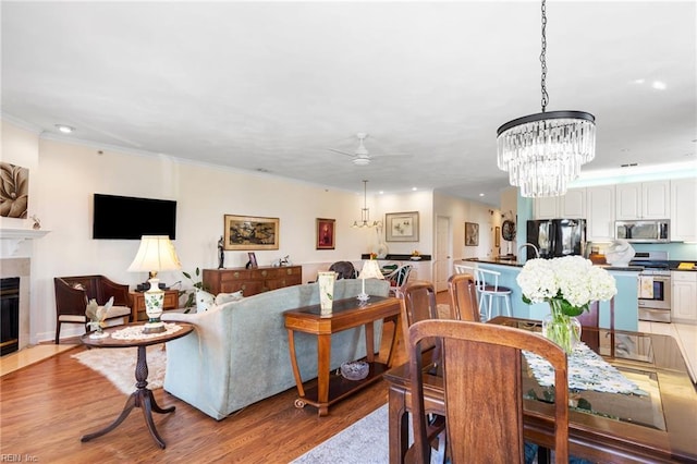 dining area featuring light wood-style floors, a tiled fireplace, ceiling fan with notable chandelier, recessed lighting, and ornamental molding