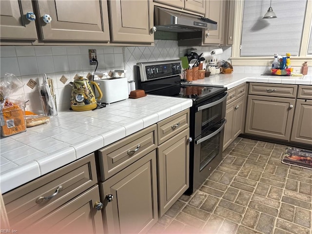 kitchen with decorative backsplash, brick patterned floor, under cabinet range hood, range with two ovens, and decorative light fixtures