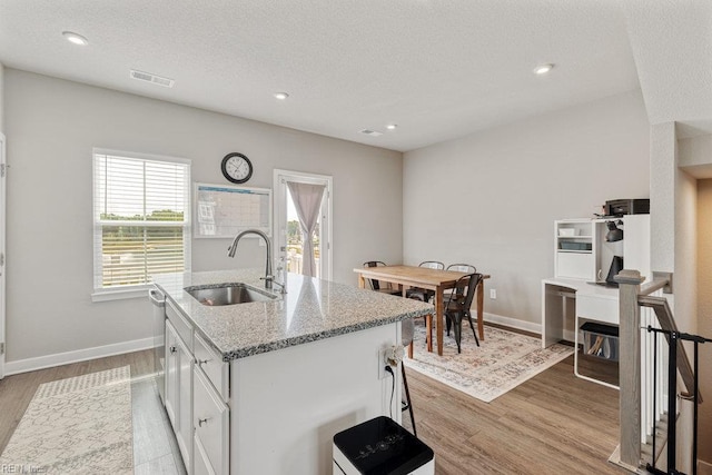 kitchen featuring light stone countertops, a sink, light wood-type flooring, visible vents, and a center island with sink