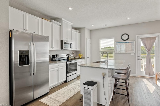 kitchen with a kitchen island with sink, white cabinets, a sink, light wood-style flooring, and appliances with stainless steel finishes