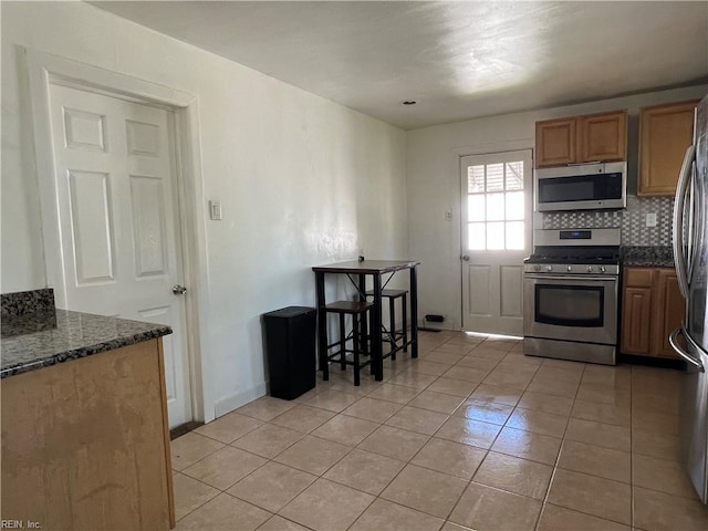 kitchen featuring decorative backsplash, brown cabinetry, appliances with stainless steel finishes, light tile patterned floors, and dark stone countertops