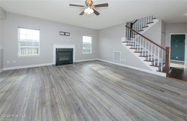 unfurnished living room with visible vents, a glass covered fireplace, wood finished floors, ceiling fan, and stairway