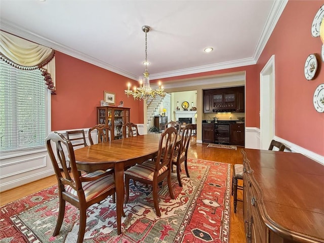 dining space featuring crown molding, a lit fireplace, light wood-type flooring, a wainscoted wall, and a chandelier