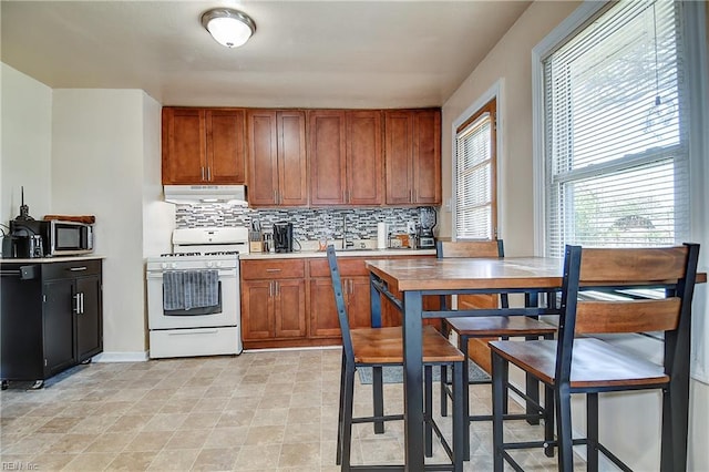 kitchen featuring backsplash, under cabinet range hood, brown cabinets, and white gas range
