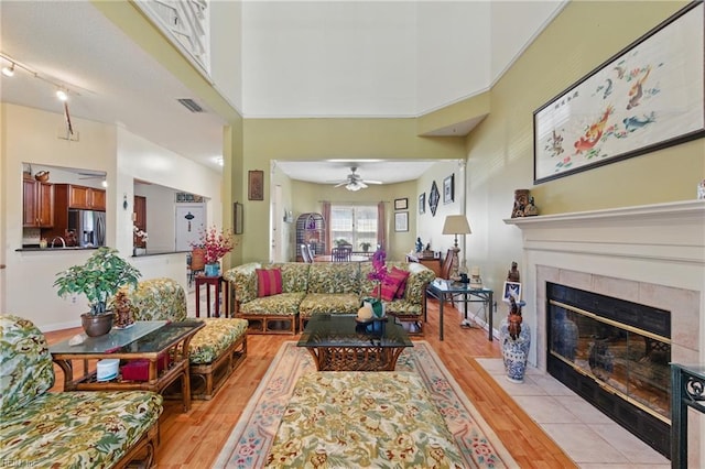 living area with visible vents, a towering ceiling, a tile fireplace, a ceiling fan, and light wood-style flooring