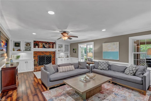 living area featuring a fireplace, built in shelves, ornamental molding, a ceiling fan, and dark wood-style floors
